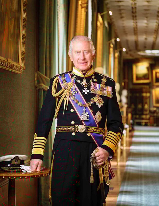 A distinguished man in an ornate military uniform with medals and sashes stands in an opulent, art-filled hallway, resting one hand on a table with a naval hat. He smiles at the camera amid luxurious surroundings.