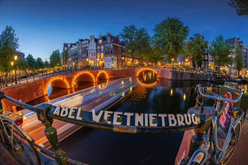 A scenic evening view of Amsterdam’s canals, with light trails from boats, illuminated arched bridges, historic buildings, and a sign reading "Abel Weetnietbrug" in the foreground.