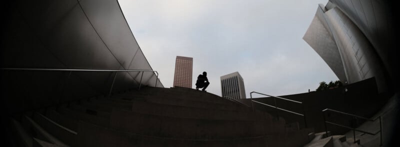 A person crouches on outdoor concrete steps surrounded by modern metal buildings and tall skyscrapers under a cloudy sky, photographed from a low angle.