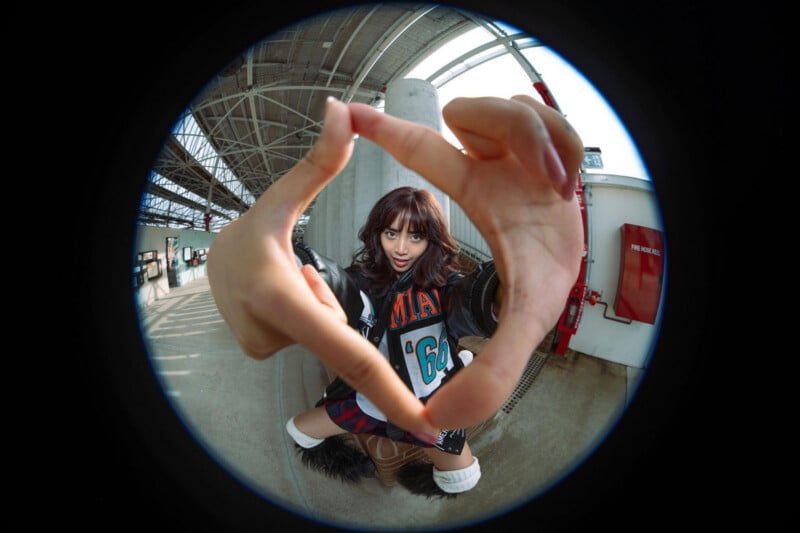 A woman poses indoors making a heart shape with her hands toward the camera, which is distorted with a fisheye lens. She wears a letterman jacket, skirt, and boots, with industrial elements in the background.