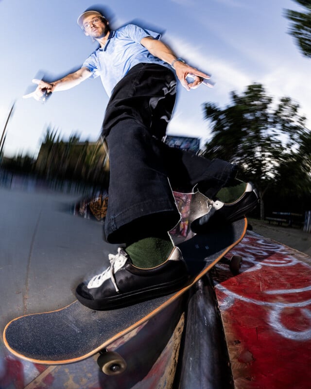 A skateboarder in a blue shirt and black pants performs a trick on a rail at a skatepark, captured from a low angle with a dynamic, wide-angle lens effect. Trees and graffiti are visible in the background.