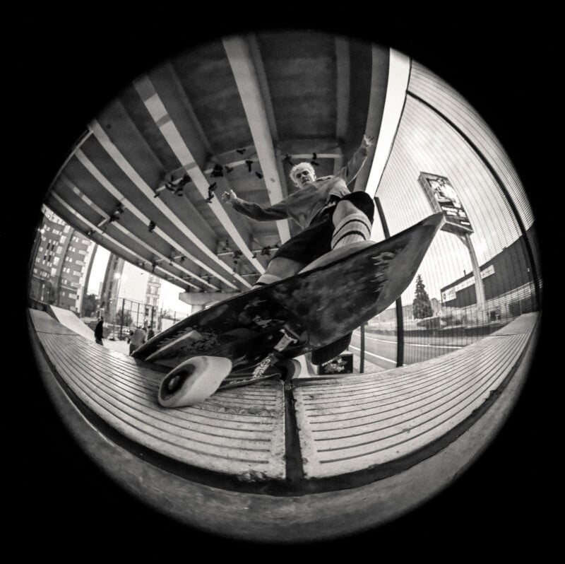 A fisheye lens shot of a skateboarder performing a trick on a ledge under an urban overpass, with tall buildings and a skatepark visible in the background. The angle emphasizes the skateboard and movement.