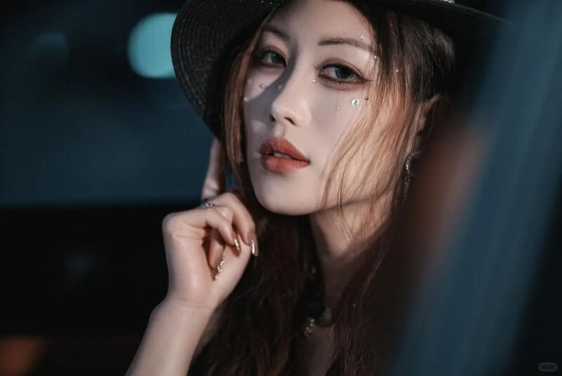 A woman with long hair wears a black hat and glittery makeup with rhinestones under her eyes. She looks directly at the camera, touching her face gently, in a dimly lit setting.