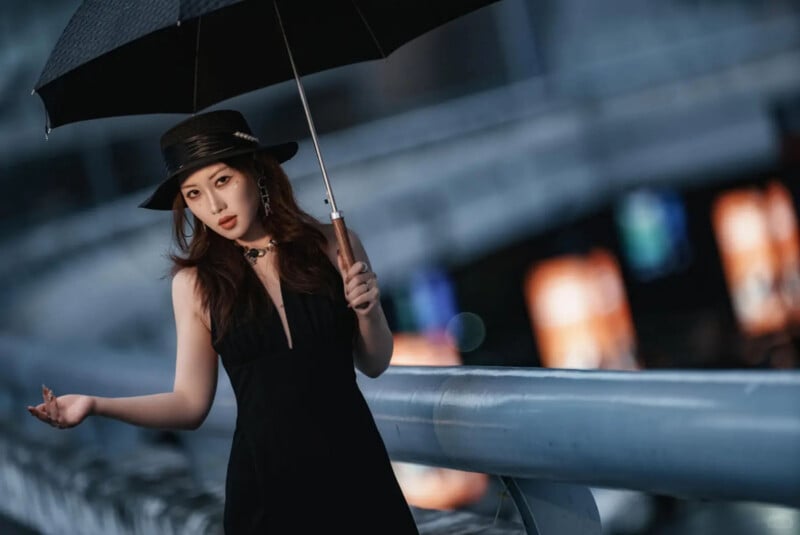 A woman in a black dress and hat stands outdoors holding a black umbrella. She leans against a metal railing with blurred city lights in the background, creating a dramatic and stylish evening scene.