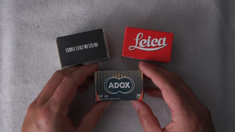 Two hands hold a box of Adox HR 50 film below two other boxes labeled "Ernst Leitz Wetzlar" and "Leica," all placed on a light gray textured surface.