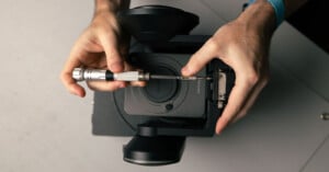 A person uses a screwdriver to repair or assemble a black camera device, viewed from above, with both hands visible holding the tool and device.