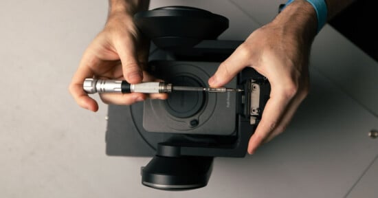 A person uses a screwdriver to repair or assemble a black camera device, viewed from above, with both hands visible holding the tool and device.