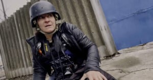 A journalist wearing a helmet and "PRESS" vest crouches on the ground near a corrugated fence and blue metal door, looking anxiously to the side while holding a camera.