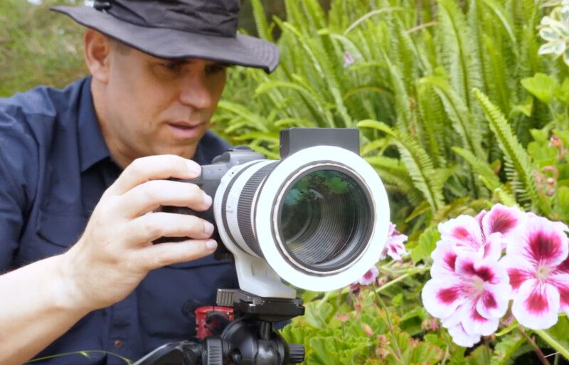 A person in a black hat and navy shirt uses a camera with a large lens on a tripod to photograph pink and white flowers in a lush garden.