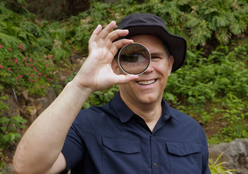 A smiling person wearing a dark hat and navy shirt holds a round glass filter up to one eye, with greenery and flowers in the background.