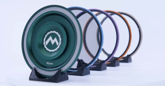 Five colorful camera lens filters, each mounted on individual black stands, are displayed in a line on a white surface. The front filter is green and shows the text "82mm" and a stylized mountain logo.