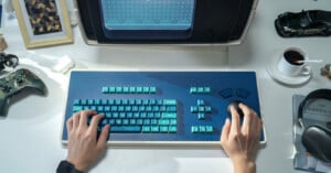 A person types on a retro-style blue keyboard with a large trackball, in front of a vintage monitor. The desk holds a cup of coffee, a book, a toy car, a framed picture, headphones, and a game controller.
