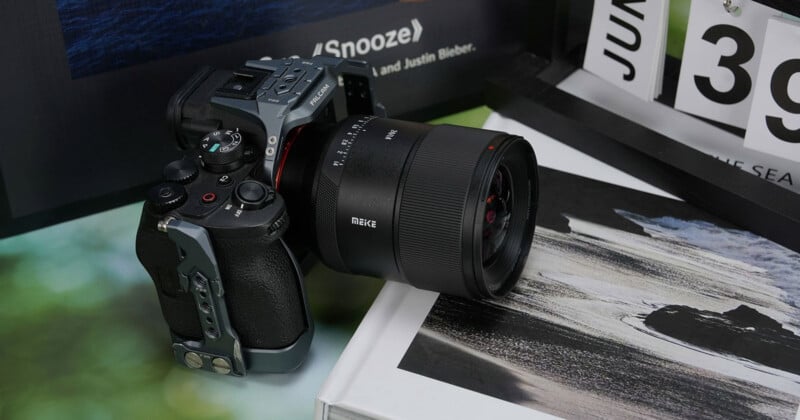 A digital camera with a large lens sits on a table next to a calendar displaying "JUN 39" and a book with a black and white cover image, with blurred green and dark backgrounds.