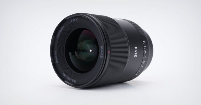 A black camera lens labeled “24/1.4” lies on its side against a plain white background, showing the glass elements and focus markings.