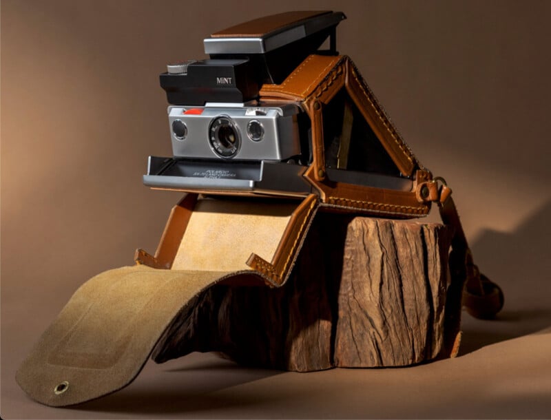 A vintage instant film camera in a brown leather case rests on a textured piece of wood, with a tan background and soft lighting highlighting the camera's metallic details.