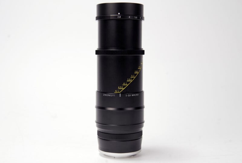 A black Zhongyi 1x-5x macro camera lens stands vertically against a plain white background, showing yellow focus markings and aperture values.