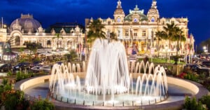 A large, illuminated fountain in front of the ornate Monte Carlo Casino at night, with palm trees, colorful flowers, and city lights in the background.