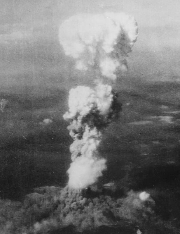 Black and white photo showing a large mushroom cloud rising high into the sky after an explosion, with dark smoke billowing above a cityscape below.