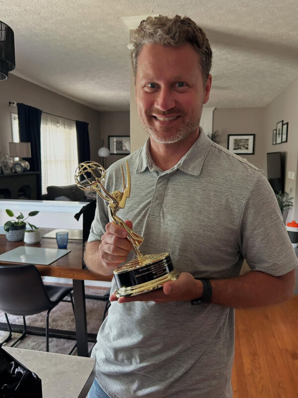A smiling man with light brown hair and a gray polo shirt holds a gold Emmy Award trophy in a modern, well-lit living room with a dining table and decor in the background.