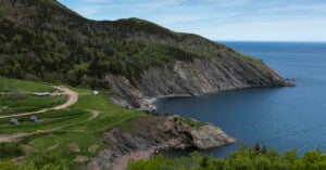 A scenic coastal landscape with steep, rocky cliffs, a winding dirt road, green hills, scattered trees, and small buildings near the edge of the ocean under a partly cloudy sky.