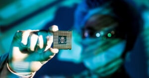 A person wearing medical gloves, a face mask, and a hair cover holds up a computer microchip, with the background blurred and blue-green lighting creating a high-tech atmosphere.