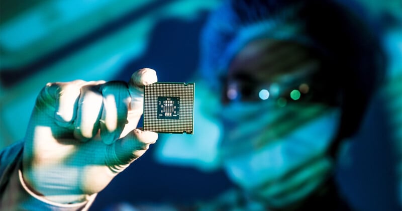 A person wearing medical gloves, a face mask, and a hair cover holds up a computer microchip, with the background blurred and blue-green lighting creating a high-tech atmosphere.