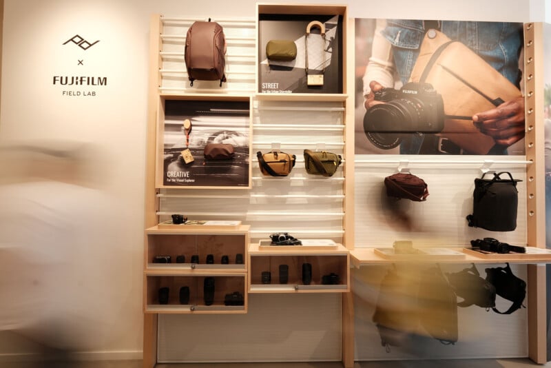 A FujiFilm Field Lab display features camera bags, lenses, and accessories on wooden shelves. Blurred figures walk by, and a large wall graphic shows hands holding a Fujifilm camera with a tan bag.