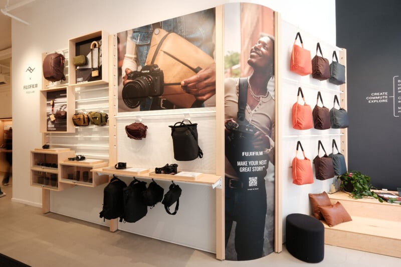 A modern store display showcases backpacks, cameras, and tote bags on wooden shelves. A large poster features a smiling woman holding a camera. The area is well-lit, minimalistic, and inviting.