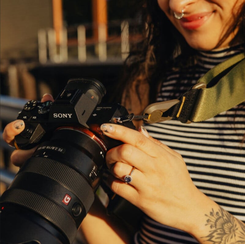 A person with a nose ring and tattoos, wearing a striped sleeveless top, smiles while holding a Sony camera outdoors in warm sunlight.