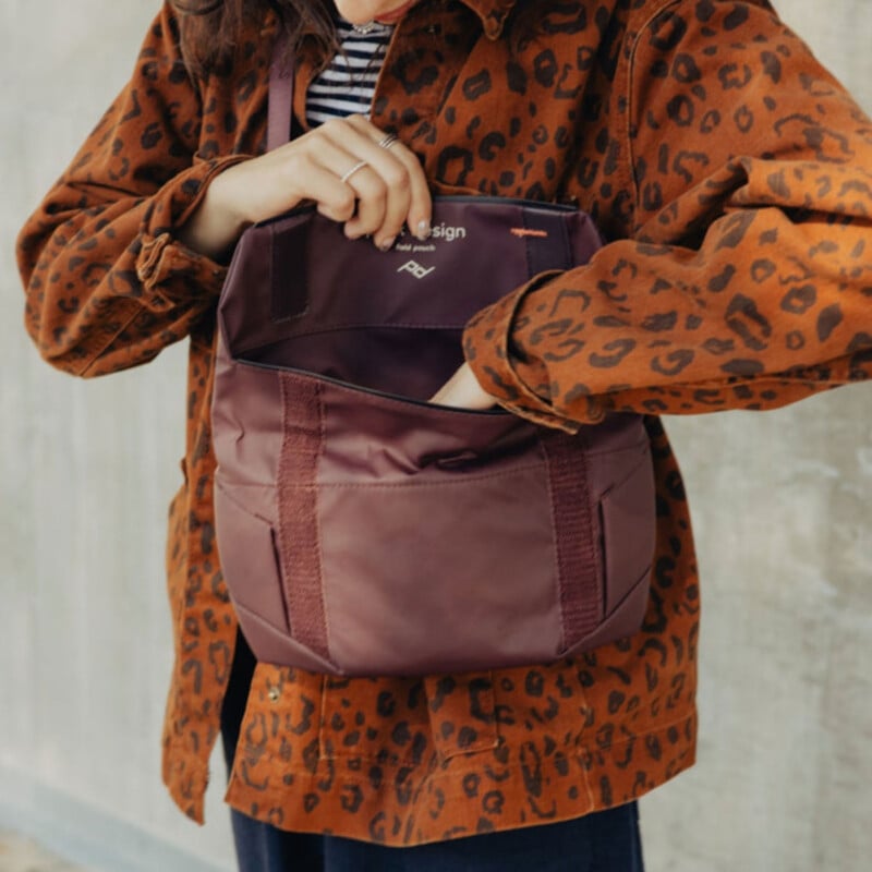 A person wearing a brown, leopard-print jacket reaches into an open, maroon-colored Peak Design pouch while standing against a gray wall.