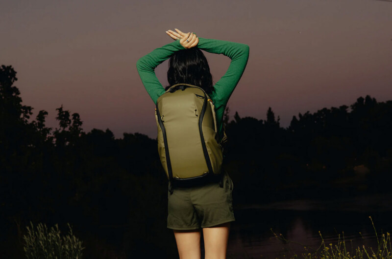 A person with short dark hair, wearing a green long-sleeve shirt, green shorts, and an olive backpack, stands outdoors at dusk with arms crossed above their head, facing away from the camera toward trees and a river.