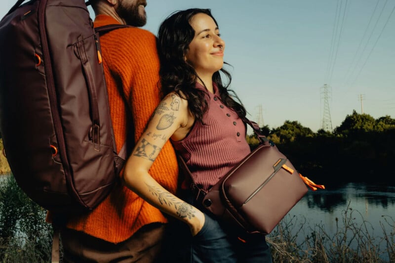 Two people stand back to back outdoors, both smiling. One carries a large backpack, and the other wears a crossbody bag. They are dressed in casual clothes, with trees and a river in the background.