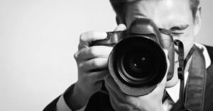 A person in a suit holding a DSLR camera up to their face, looking through the viewfinder and preparing to take a photo. The image is in black and white.