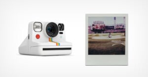 A white instant camera with rainbow stripes is shown next to a developed instant photo of a baseball game on a stadium field. The camera and photo are on a plain white background.