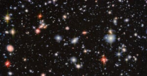 A deep space image showing countless distant galaxies of various sizes and colors scattered across a black background, with some bright, colorful stars featuring visible diffraction spikes.