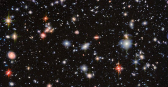 A deep space image showing countless distant galaxies of various sizes and colors scattered across a black background, with some bright, colorful stars featuring visible diffraction spikes.