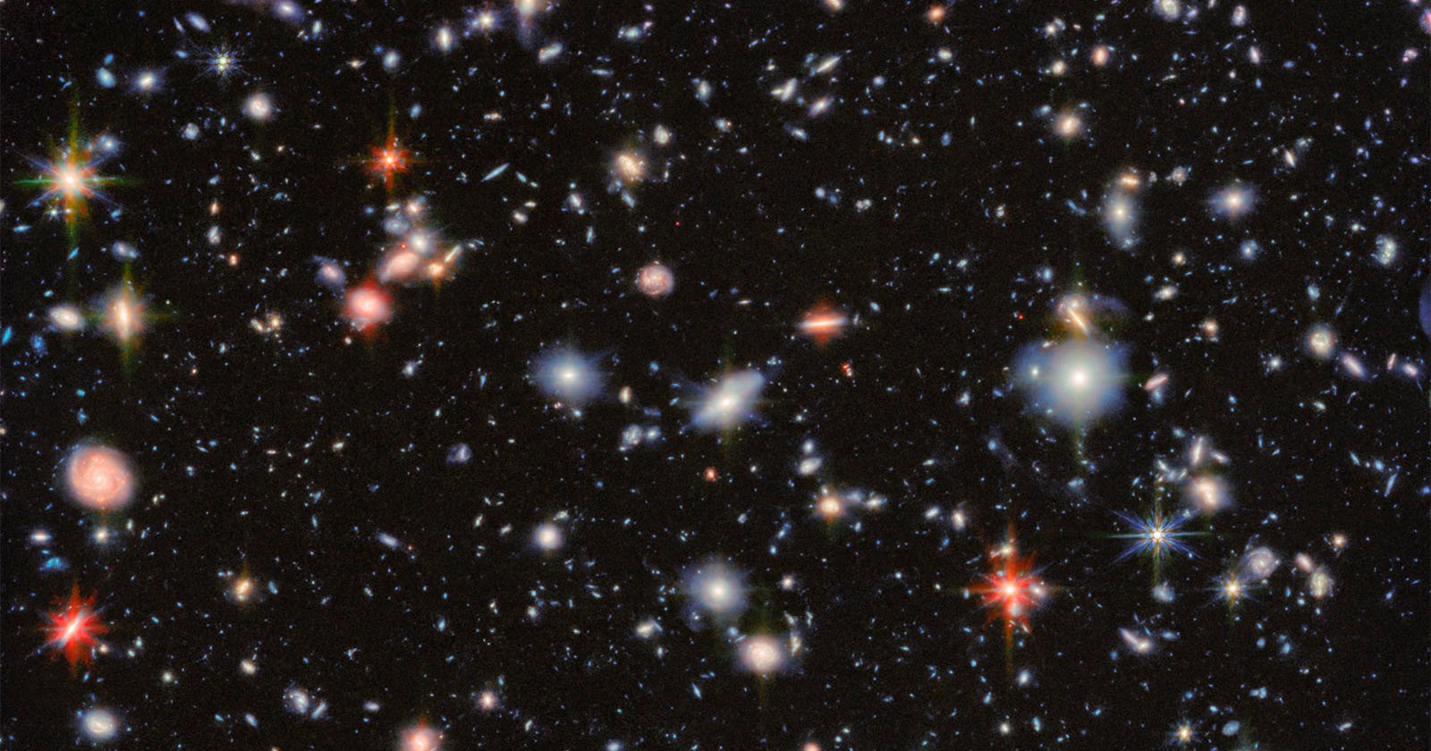 A deep space image showing countless distant galaxies of various sizes and colors scattered across a black background, with some bright, colorful stars featuring visible diffraction spikes.