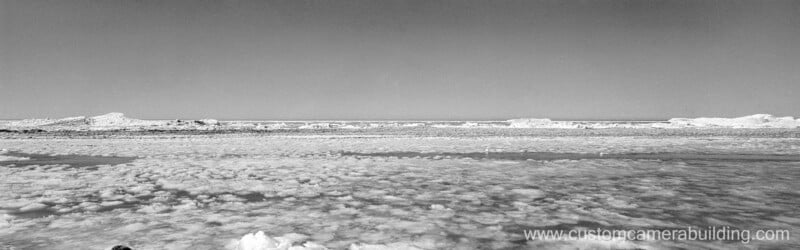 Wide view of a frozen, icy landscape with scattered snow and ice formations under a clear sky. The scene appears cold and desolate. The website "www.customcamerabuilding.com" is visible in the lower right corner.