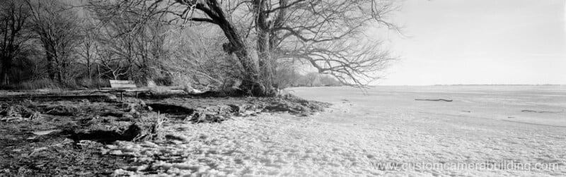 Panoramic black-and-white photo of a snowy shoreline with bare trees and a wooden bench on the left, looking out over a frozen lake under a clear sky.