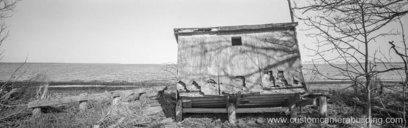 An old, weathered wooden shack stands on a barren, overgrown shoreline near a body of water, with leafless trees and broken benches in the foreground. The website "www.customcamerabuilding.com" is visible.