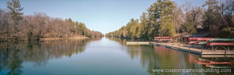 A calm river flanked by trees on both sides, with docks and stacked red canoes on the right. The sky is clear, and the water reflects the surrounding landscape. Website www.customcamerabuilding.com appears at the bottom right.