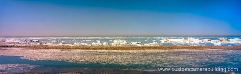 Wide panoramic view of a frozen shoreline with patches of ice and snow along the water under a clear blue sky; the website www.customcamerabuilding.com appears in the bottom right corner.