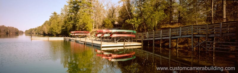 A lakeside dock with stacked canoes and a wooden ramp, surrounded by trees reflected in calm water; the website www.customcamerabuilding.com is visible in the bottom right corner.