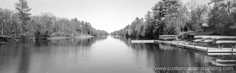 A calm river lined with leafless and evergreen trees, with a dock and boats on the right side. Reflections of trees appear on the water. Website "www.customcamerabuilding.com" is at the bottom right.