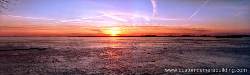 A vibrant sunset over a calm body of water with colorful clouds and contrails in the sky; the sun is low on the horizon and the website "www.customcamerabuilding.com" appears in the lower right corner.