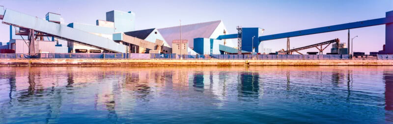 A modern industrial facility with blue geometric buildings and conveyor belts is reflected in calm water under a clear blue sky.