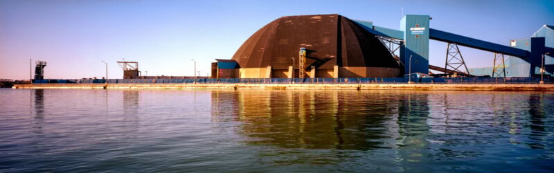 A large industrial facility with a black dome building and blue structures stands on the waterfront, reflecting in the calm water under a clear sky.