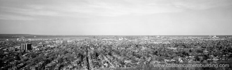 Black and white panoramic view of a city with dense residential areas, taller buildings, a river, and industrial structures in the distance; website "www.customcamerabuilding.com" appears in the bottom right.