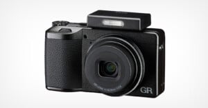 A compact black digital camera with a textured grip, extended lens, and raised rectangular pop-up flash. The letters "GR" are visible on the lower right corner of the camera front.