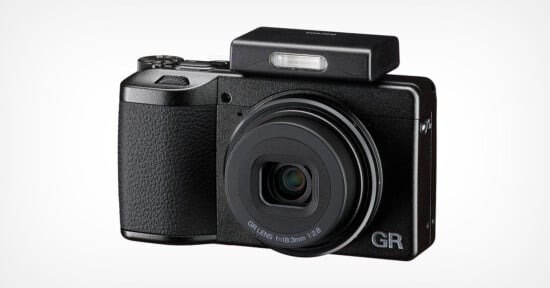 A compact black digital camera with a textured grip, extended lens, and raised rectangular pop-up flash. The letters "GR" are visible on the lower right corner of the camera front.